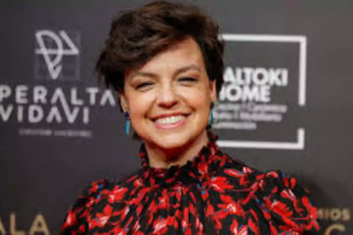 Mujer de cabello corto y oscuro sonríe frente a un fondo con logotipos vistiendo una blusa de estampado rojo y negro Mujer de cabello corto y oscuro sonríe frente a un fondo con logotipos vistiendo una blusa de estampado rojo y negro