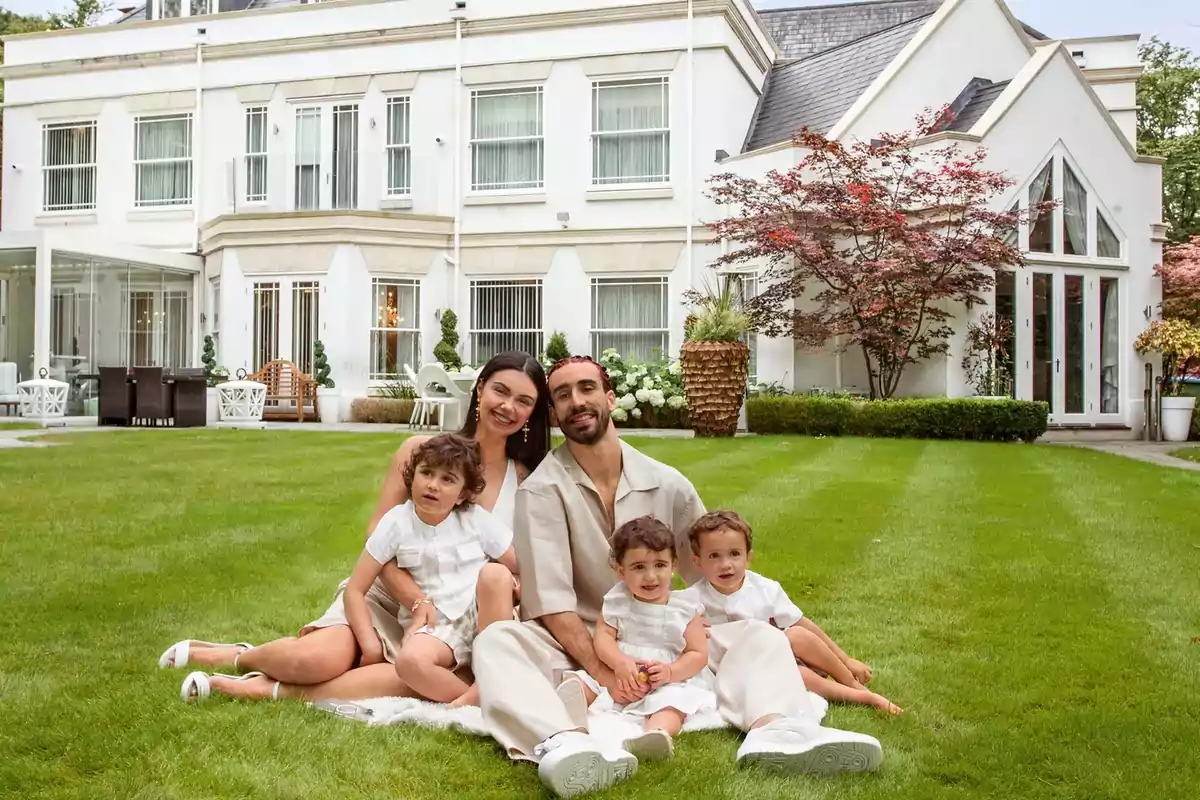 Família de cinc persones asseguda sobre la gespa davant d’una casa gran i elegant gaudint d’un dia a l’aire lliure. Família de cinc persones asseguda sobre la gespa davant d’una casa gran i elegant gaudint d’un dia a l’aire lliure.