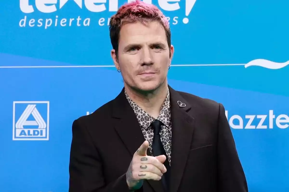 Dani Martín, vestido con traje negro y camisa estampada, señalando hacia el frente sobre un fondo azul con logotipos. Dani Martín, vestido con traje negro y camisa estampada, señalando hacia el frente sobre un fondo azul con logotipos.