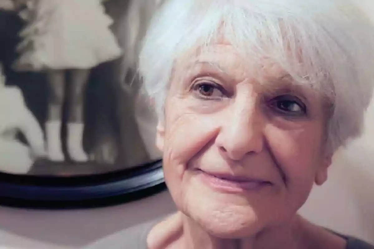 Dona gran de cabells blancs mirant cap a un costat amb una expressió serena i un retrat antic en blanc i negre al fons Dona gran de cabells blancs mirant cap a un costat amb una expressió serena i un retrat antic en blanc i negre al fons