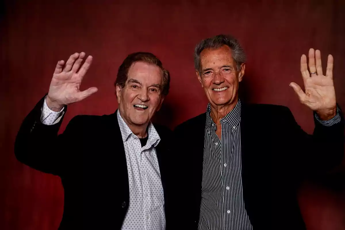 El Dúo Dinámico sonríen y saludan con la mano levantada frente a un fondo rojo, ambos visten camisas y sacos oscuros El Dúo Dinámico sonríen y saludan con la mano levantada frente a un fondo rojo, ambos visten camisas y sacos oscuros