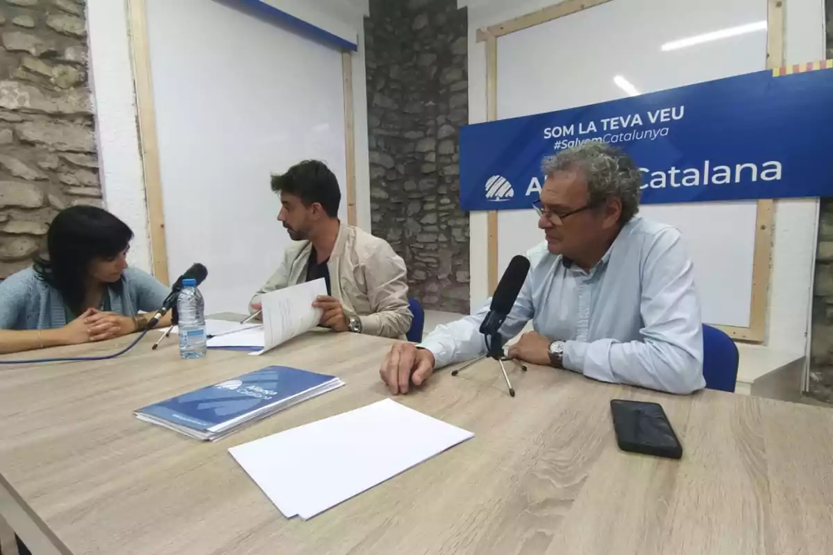 Tres persones assegudes en una taula de fusta conversen davant de micròfons i documents, amb un cartell blau de fons que diu Tres persones assegudes en una taula de fusta conversen davant de micròfons i documents, amb un cartell blau de fons que diu