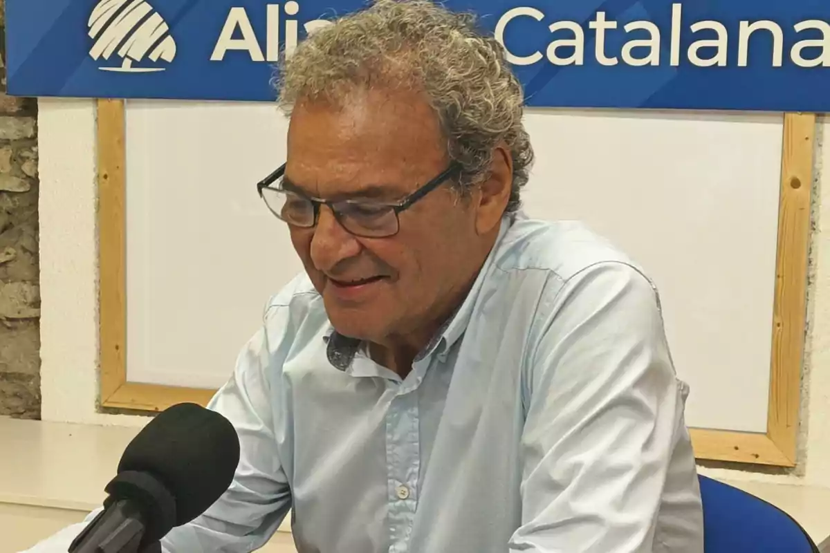 Home de cabells canosos i ulleres parlant davant d’un micròfon en un estudi amb un cartell blau de fons Home de cabells canosos i ulleres parlant davant d’un micròfon en un estudi amb un cartell blau de fons