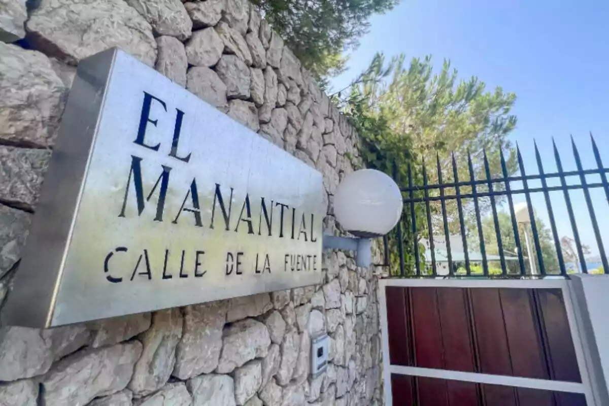 Rètol metàl·lic amb el text El Manantial Carrer de la Font en una paret de pedra al costat d’una reixa i vegetació al fons Rètol metàl·lic amb el text El Manantial Carrer de la Font en una paret de pedra al costat d’una reixa i vegetació al fons