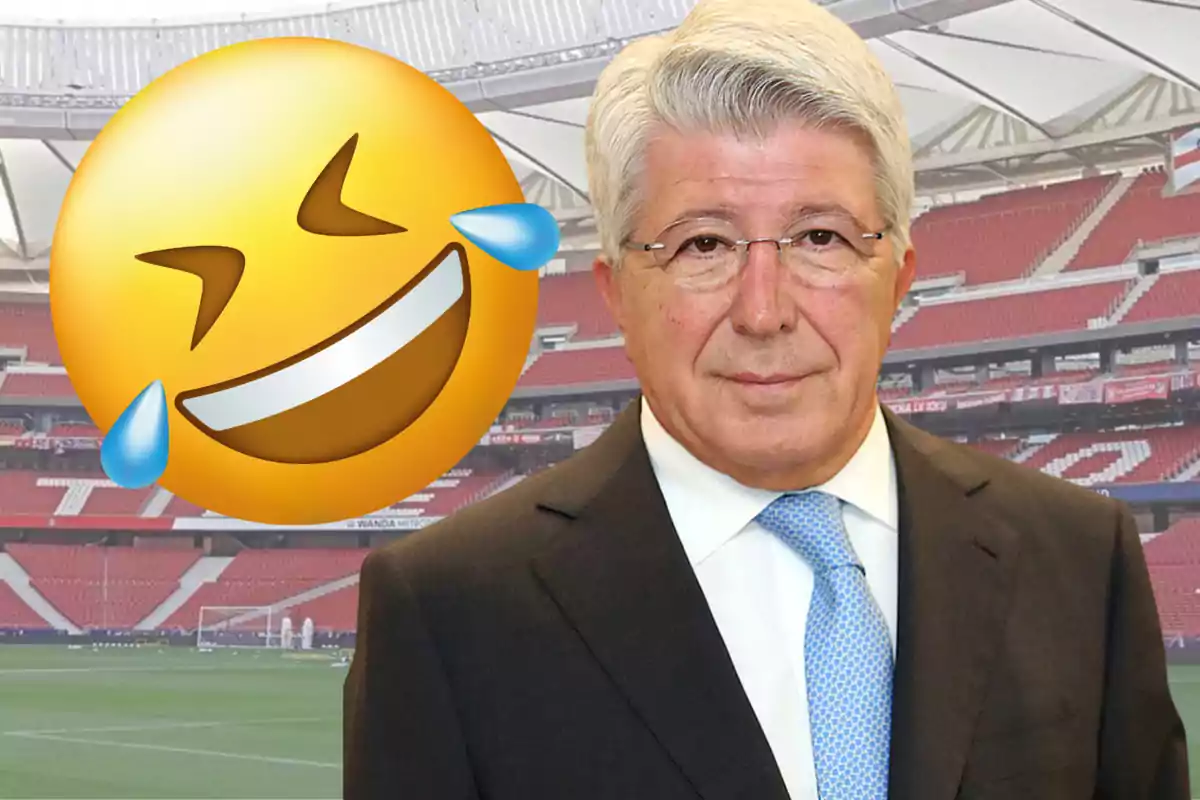 Hombre de cabello canoso con traje y corbata azul posando en un estadio de fútbol junto a un emoji grande de risa con lágrimas