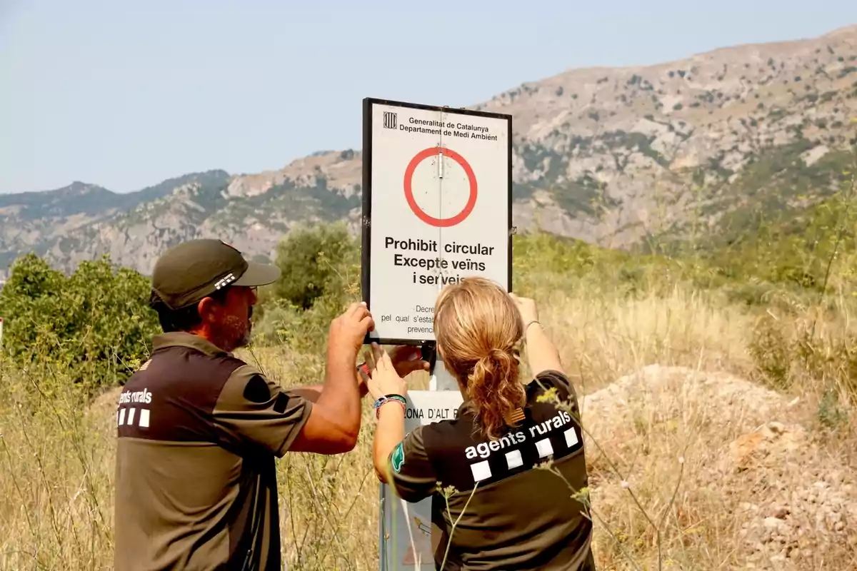 Dos agents rurals col·loquen un senyal de prohibit circular en un camí de muntanya envoltat de vegetació i turons al fons.