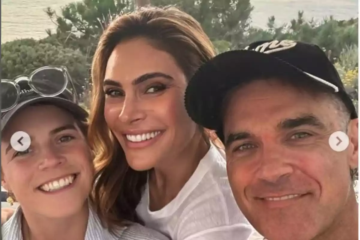 Eugenie of York, Ayda Field Williams, and Robbie Williams pose smiling with vegetation and the sea behind them. Eugenie of York, Ayda Field Williams, and Robbie Williams pose smiling with vegetation and the sea behind them.