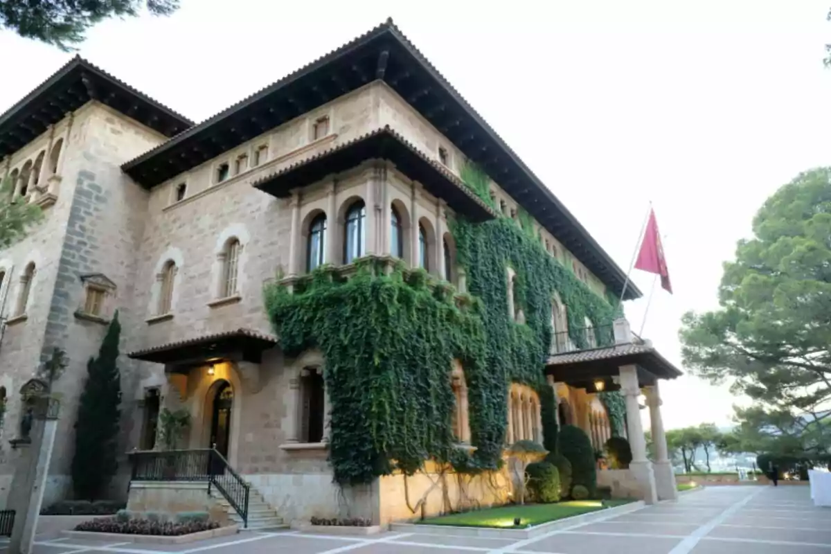 Edificio antiguo de piedra con ventanas arqueadas y enredaderas verdes cubriendo parte de la fachada, una bandera roja ondea en la entrada y hay árboles alrededor Edificio antiguo de piedra con ventanas arqueadas y enredaderas verdes cubriendo parte de la fachada, una bandera roja ondea en la entrada y hay árboles alrededor