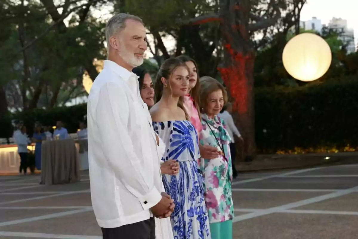 Five people pose together outdoors during an elegant event, surrounded by trees and decorative lights, all dressed in formal summer attire. Five people pose together outdoors during an elegant event, surrounded by trees and decorative lights, all dressed in formal summer attire.