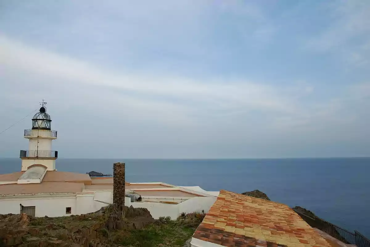 Far blanc vora el mar amb cel serè i teulada de teules en primer pla Far blanc vora el mar amb cel serè i teulada de teules en primer pla
