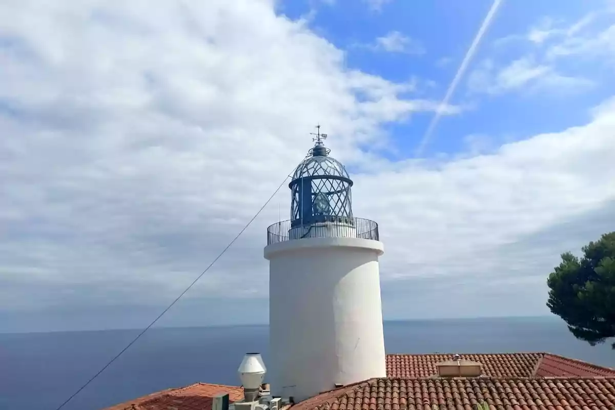 Far blanc vora el mar amb cel parcialment ennuvolat i teulada de teules vermelles en primer pla Far blanc vora el mar amb cel parcialment ennuvolat i teulada de teules vermelles en primer pla