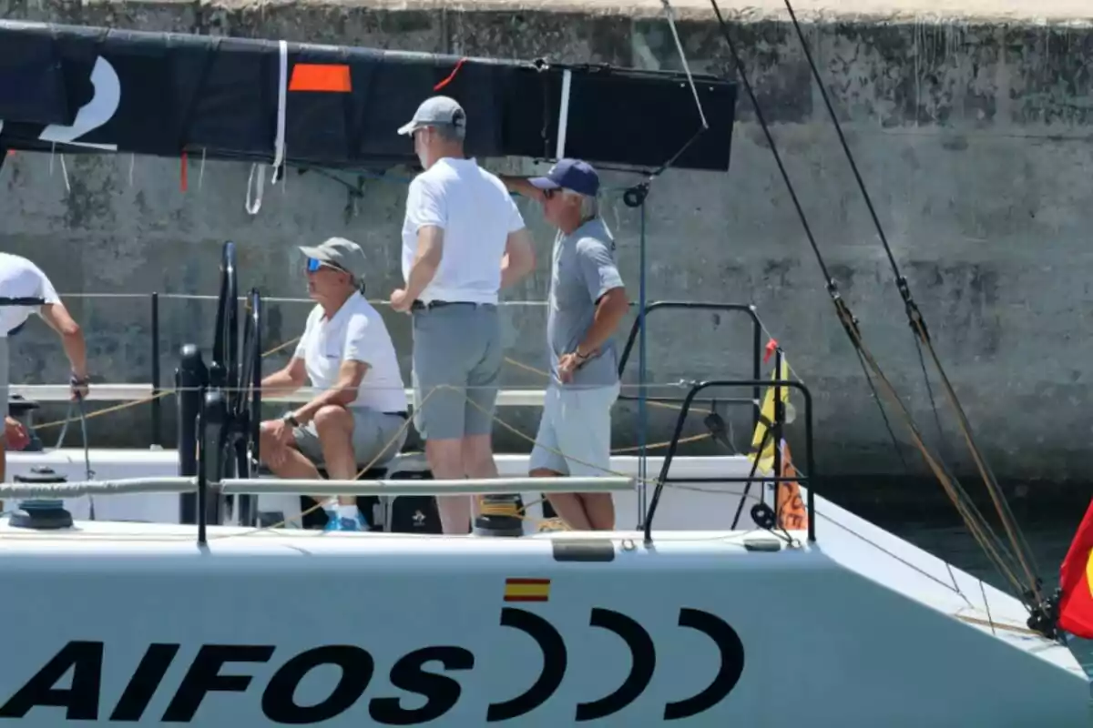 Jaime Rodríguez Toubes, Felip VI i un altre home amb roba esportiva i gorres són a bord d’un veler anomenat Aifos al costat d’un moll de pedra Jaime Rodríguez Toubes, Felip VI i un altre home amb roba esportiva i gorres són a bord d’un veler anomenat Aifos al costat d’un moll de pedra