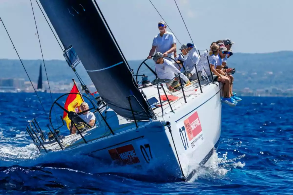 Un veler de competició navega a gran velocitat amb diversos tripulants a bord, onejant una bandera d’Espanya sobre aigües blaves i sota un cel serè. Un veler de competició navega a gran velocitat amb diversos tripulants a bord, onejant una bandera d’Espanya sobre aigües blaves i sota un cel serè.