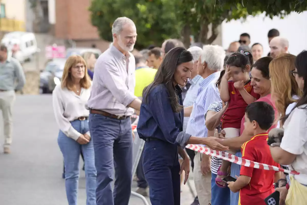 La reina Letizia sonriente saluda a un grupo de personas detrás de una cinta de seguridad en un evento al aire libre, mientras Felipe VI y más asistentes observan la escena. La reina Letizia sonriente saluda a un grupo de personas detrás de una cinta de seguridad en un evento al aire libre, mientras Felipe VI y más asistentes observan la escena.