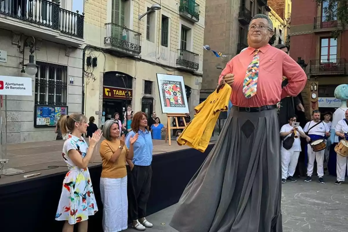 Un grup de persones aplaudeix davant d’una figura gegant vestida amb camisa rosa i corbata colorida en una plaça urbana durant un esdeveniment festiu.