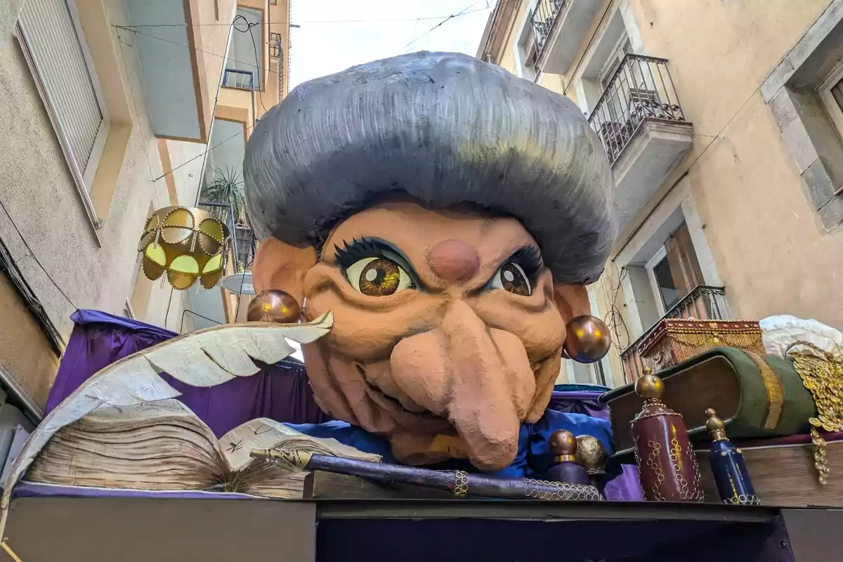 Cap gegant d’una figura caricaturesca amb un nas gros i cabells grisos, envoltat d’objectes decoratius com llibres, plomes i flascons en un carrer estret entre edificis. Cap gegant d’una figura caricaturesca amb un nas gros i cabells grisos, envoltat d’objectes decoratius com llibres, plomes i flascons en un carrer estret entre edificis.