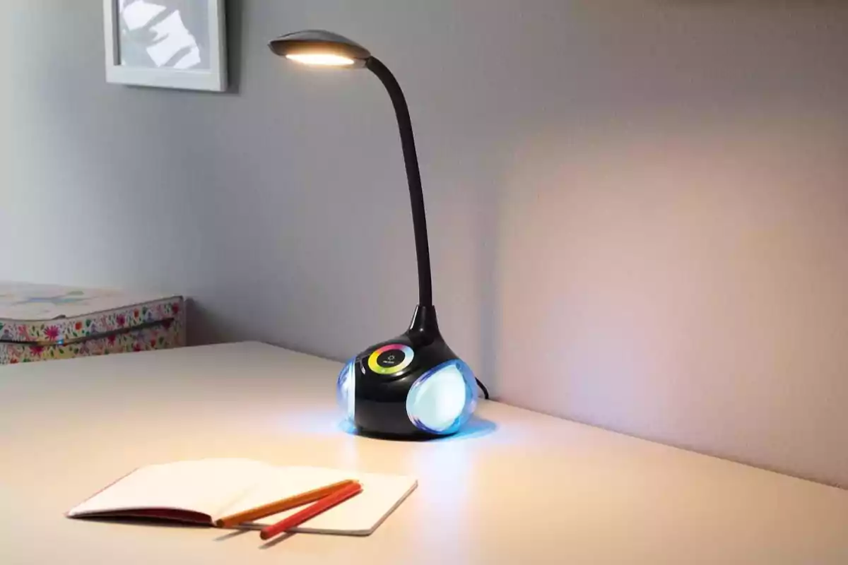 Làmpada d’escriptori moderna amb llum LED encesa sobre una taula blanca al costat d’un quadern obert i dos llapis.