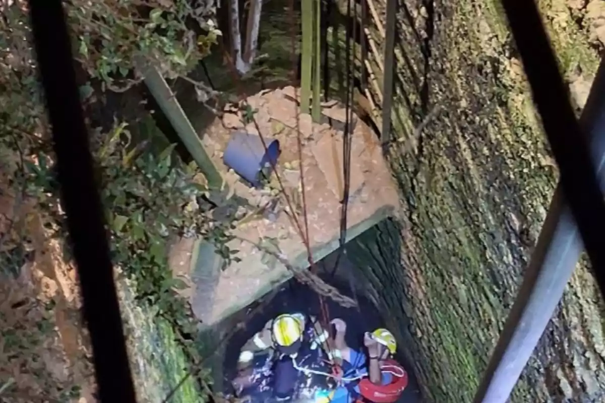 Dos rescatadors amb casc i armilla ajuden una persona dins d’un pou profund amb parets de pedra i aigua al fons, mentre cordes pengen des de la superfície.