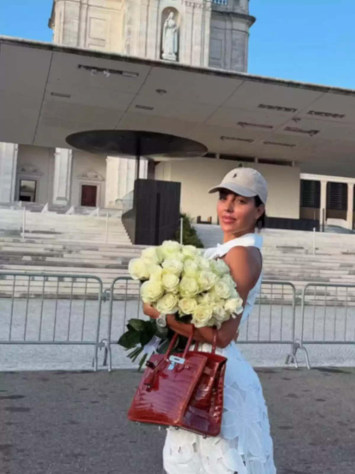 Mujer vestida de blanco con gorra clara sosteniendo un ramo de rosas blancas y un bolso rojo frente a un edificio religioso Mujer vestida de blanco con gorra clara sosteniendo un ramo de rosas blancas y un bolso rojo frente a un edificio religioso
