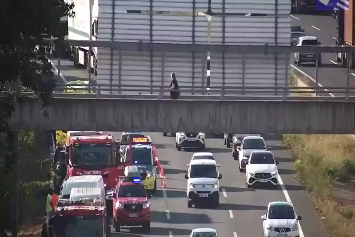 Persona caminando sobre un puente peatonal mientras varios vehículos de emergencia y autos están detenidos en una carretera Persona caminando sobre un puente peatonal mientras varios vehículos de emergencia y autos están detenidos en una carretera