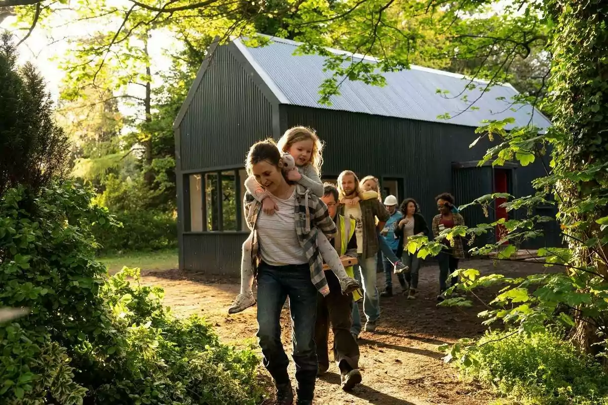Un grup de persones camina per un camí al bosc davant d’una casa moderna de color fosc, una dona porta una nena a coll mentre la resta els segueix entre arbres i vegetació sota la llum del sol. Un grup de persones camina per un camí al bosc davant d’una casa moderna de color fosc, una dona porta una nena a coll mentre la resta els segueix entre arbres i vegetació sota la llum del sol.