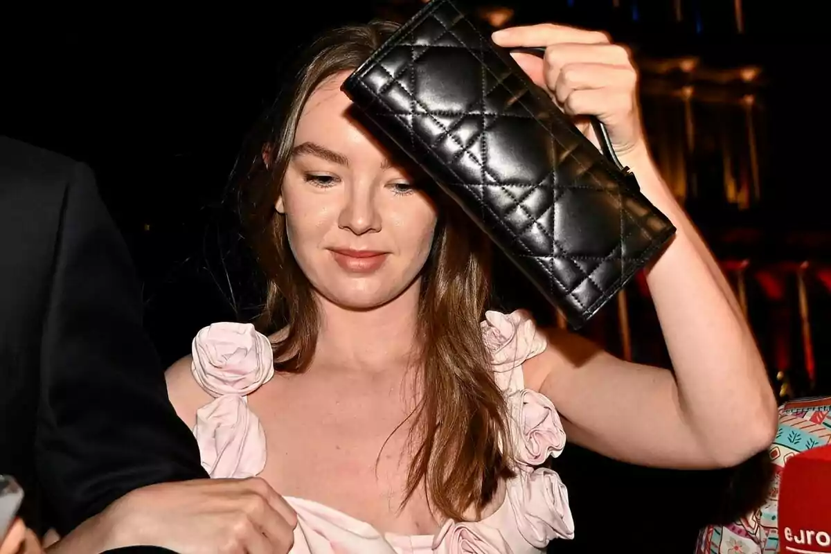 Woman in a light pink dress with floral details holds a black quilted handbag in front of her face at a nighttime event Woman in a light pink dress with floral details holds a black quilted handbag in front of her face at a nighttime event