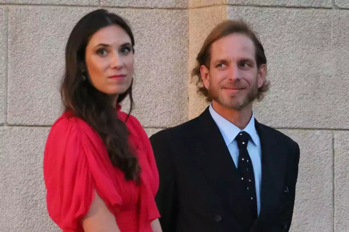 A woman in a red dress and a man in a dark suit pose together in front of a stone wall. A woman in a red dress and a man in a dark suit pose together in front of a stone wall.