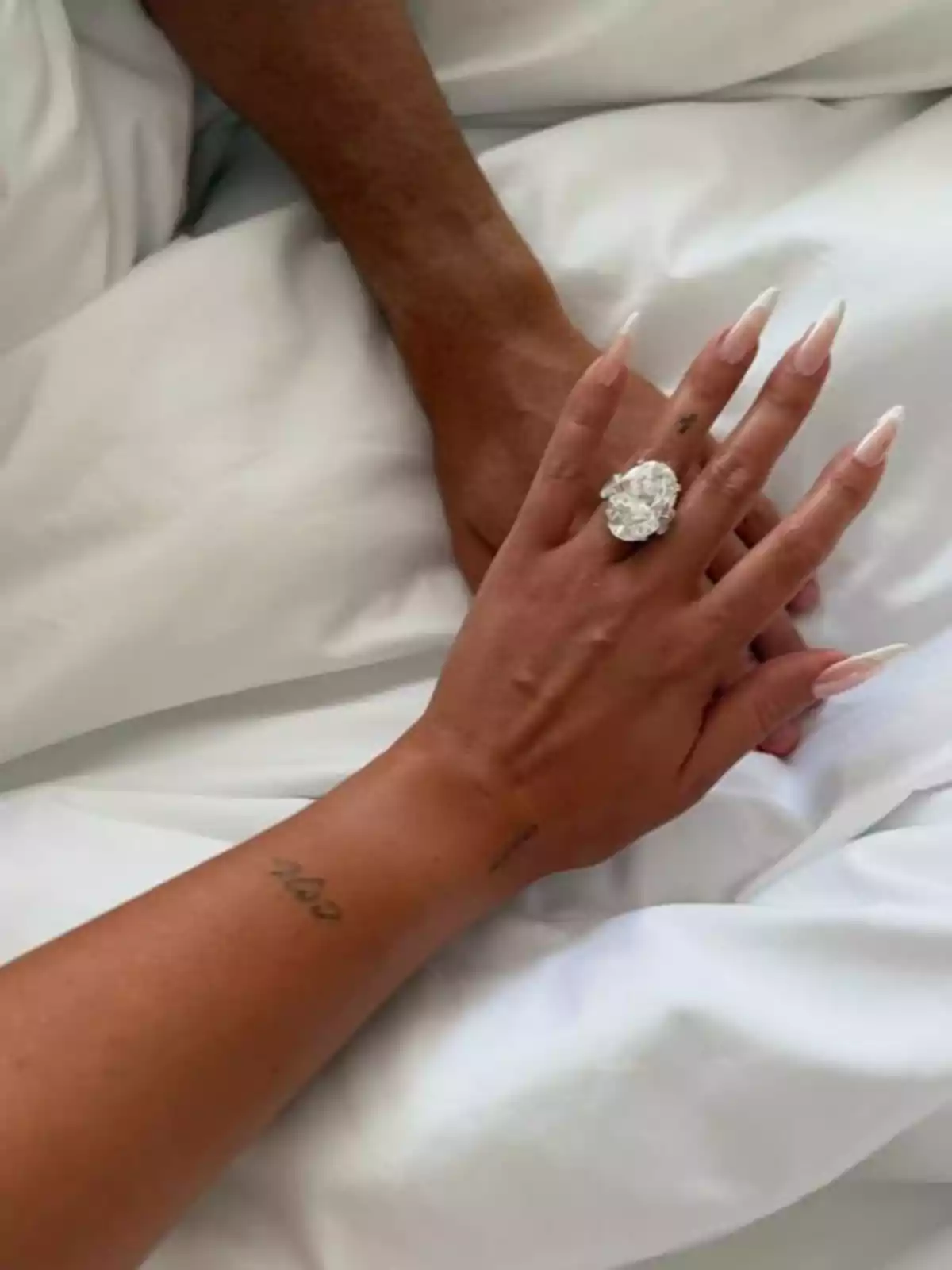 Manos entrelazadas sobre sábanas blancas, una de ellas con un anillo grande y uñas largas decoradas Manos entrelazadas sobre sábanas blancas, una de ellas con un anillo grande y uñas largas decoradas