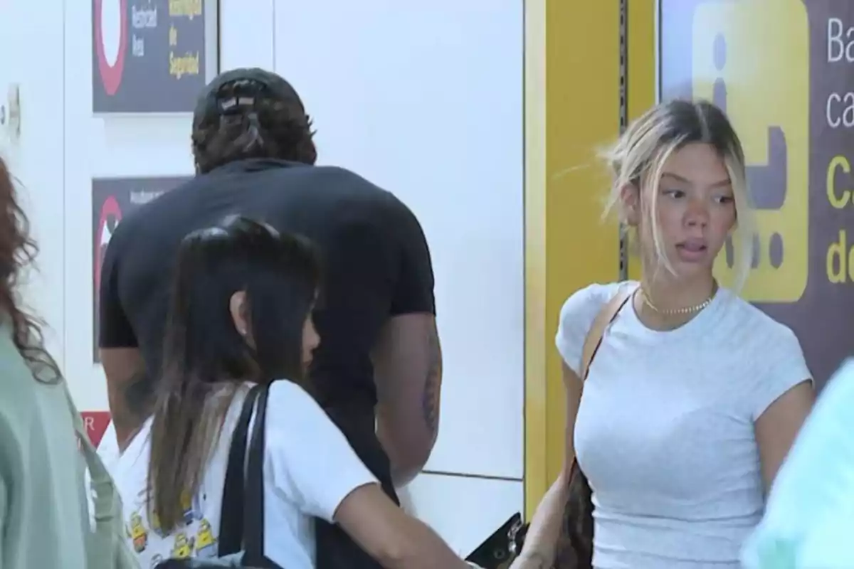 Tres persones en un aeroport, una dona rossa amb samarreta blanca mira cap a un costat mentre les altres dues persones estan d’esquena. Tres persones en un aeroport, una dona rossa amb samarreta blanca mira cap a un costat mentre les altres dues persones estan d’esquena.