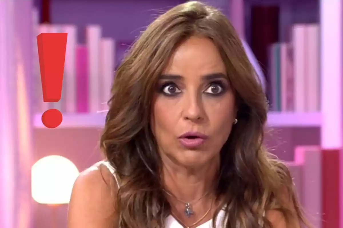 Carmen Alcayde con expresión de sorpresa y un signo de exclamación rojo a su lado en un ambiente interior con estantería de libros al fondo