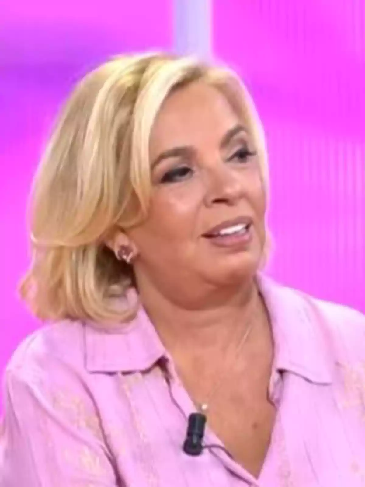 Carmen Borrego rubia con cabello corto y ondulado viste una blusa rosa y lleva un micrófono de solapa mientras sonríe en un set de televisión con fondo rosa Carmen Borrego rubia con cabello corto y ondulado viste una blusa rosa y lleva un micrófono de solapa mientras sonríe en un set de televisión con fondo rosa