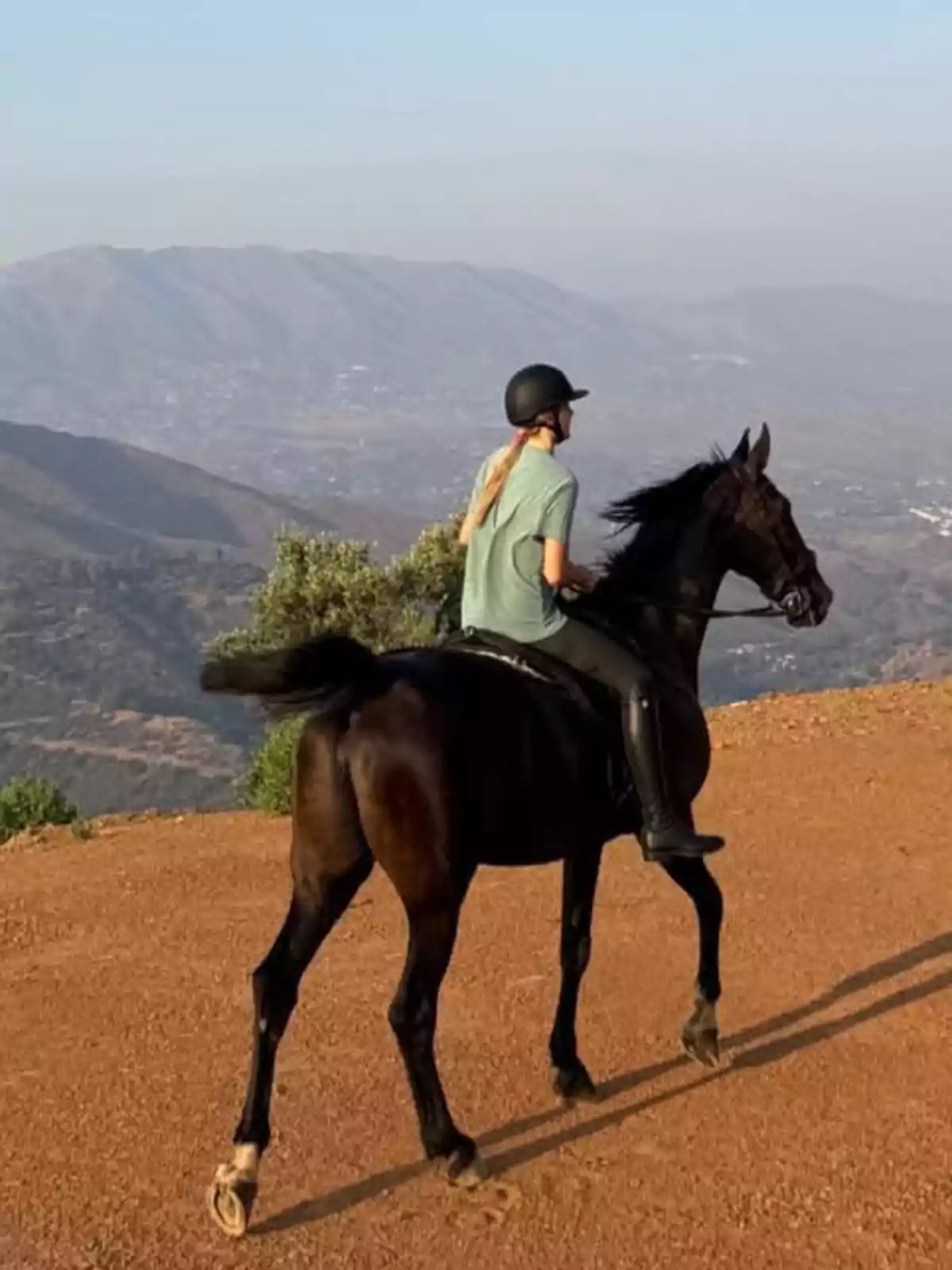 Cristina Iglesias montando un caballo negro en un sendero de tierra con montañas al fondo Cristina Iglesias montando un caballo negro en un sendero de tierra con montañas al fondo