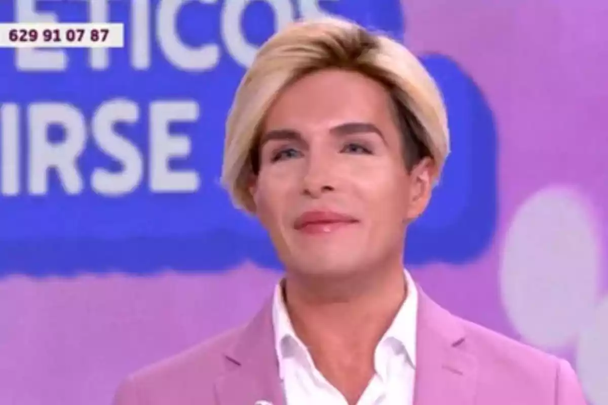 Persona de cabell ros i curt vestint una jaqueta rosa clar i una camisa blanca en un plató de televisió amb un fons morat i blau desenfocat