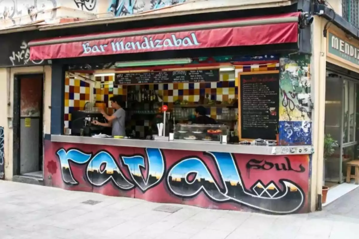 Bar de cantonada amb façana decorada amb grafit colorit que diu raval, una barra oberta a l’exterior i dues persones treballant a dins, amb rajoles de colors a la paret i menús escrits en pissarres. Bar de cantonada amb façana decorada amb grafit colorit que diu raval, una barra oberta a l’exterior i dues persones treballant a dins, amb rajoles de colors a la paret i menús escrits en pissarres.