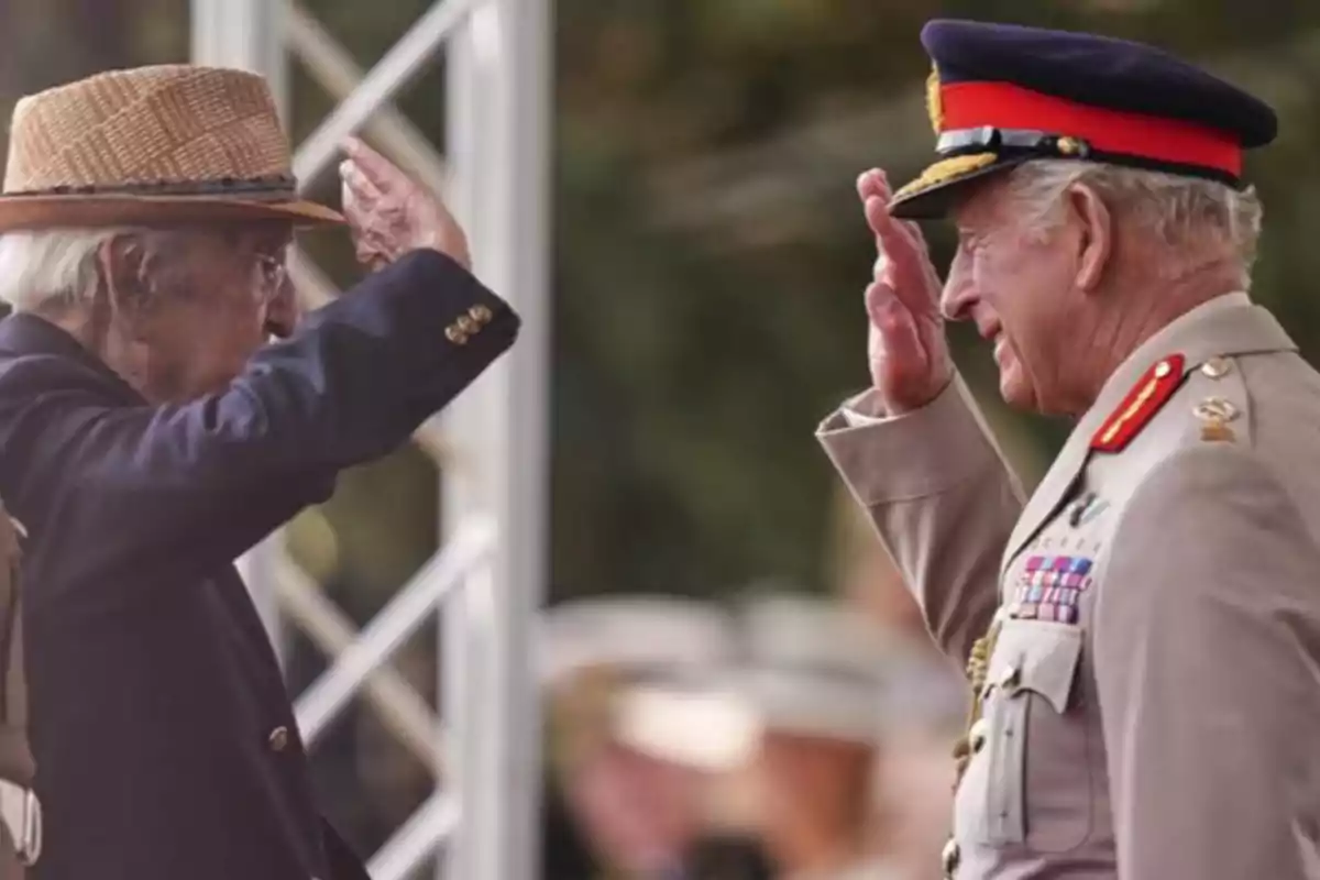 El rey Carlos III y un señor mayor se saludan militarmente uno lleva uniforme y gorra militar mientras el otro viste un saco azul y sombrero de paja