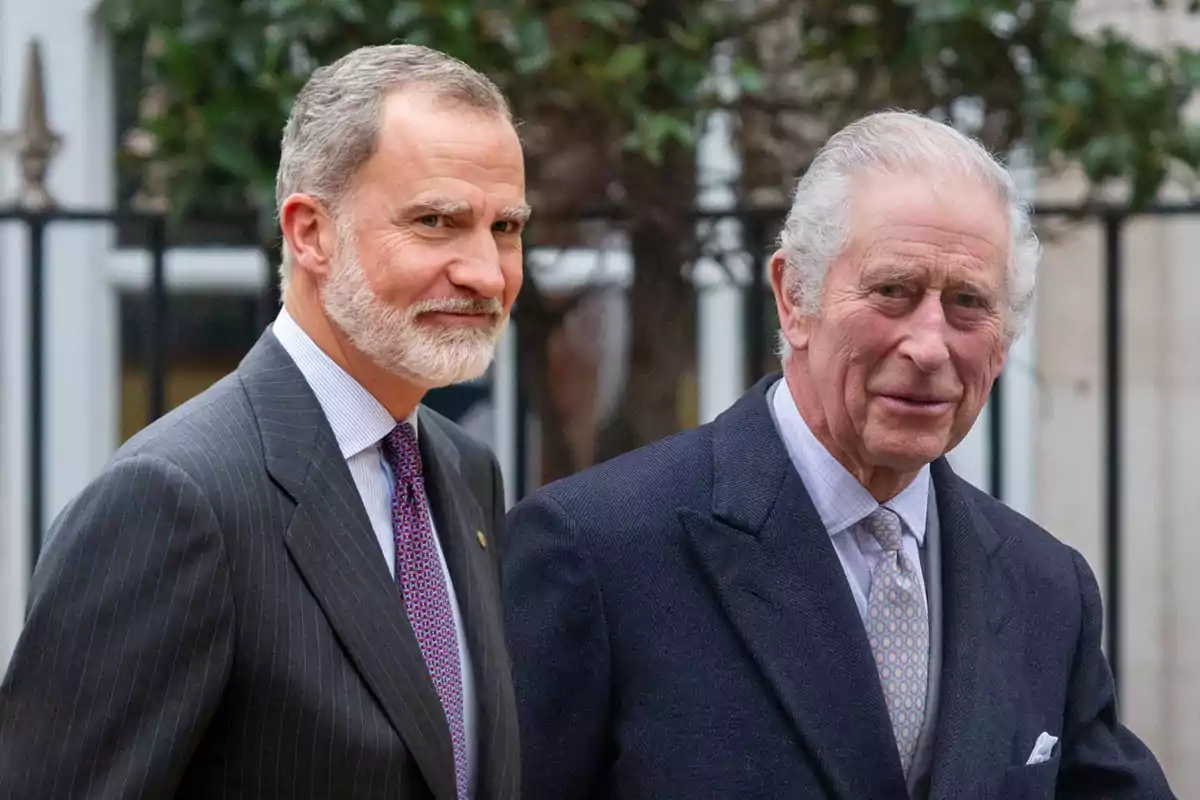 El rey Felipe VI y el rey Carlos III mayores vestidos con traje y corbata posan juntos al aire libre frente a una reja y árboles.