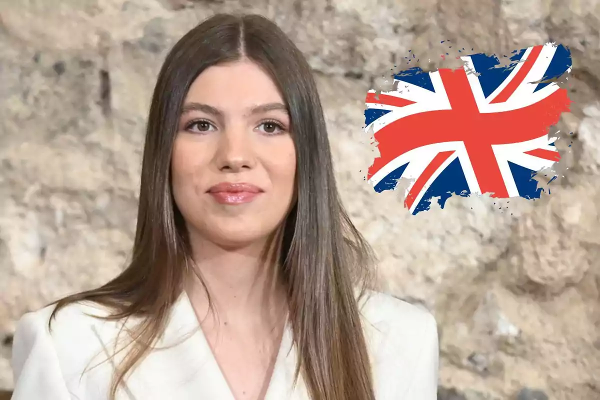 Jove de cabell llarg i llis amb jaqueta blanca davant d’una paret de pedra i una bandera del Regne Unit al fons
