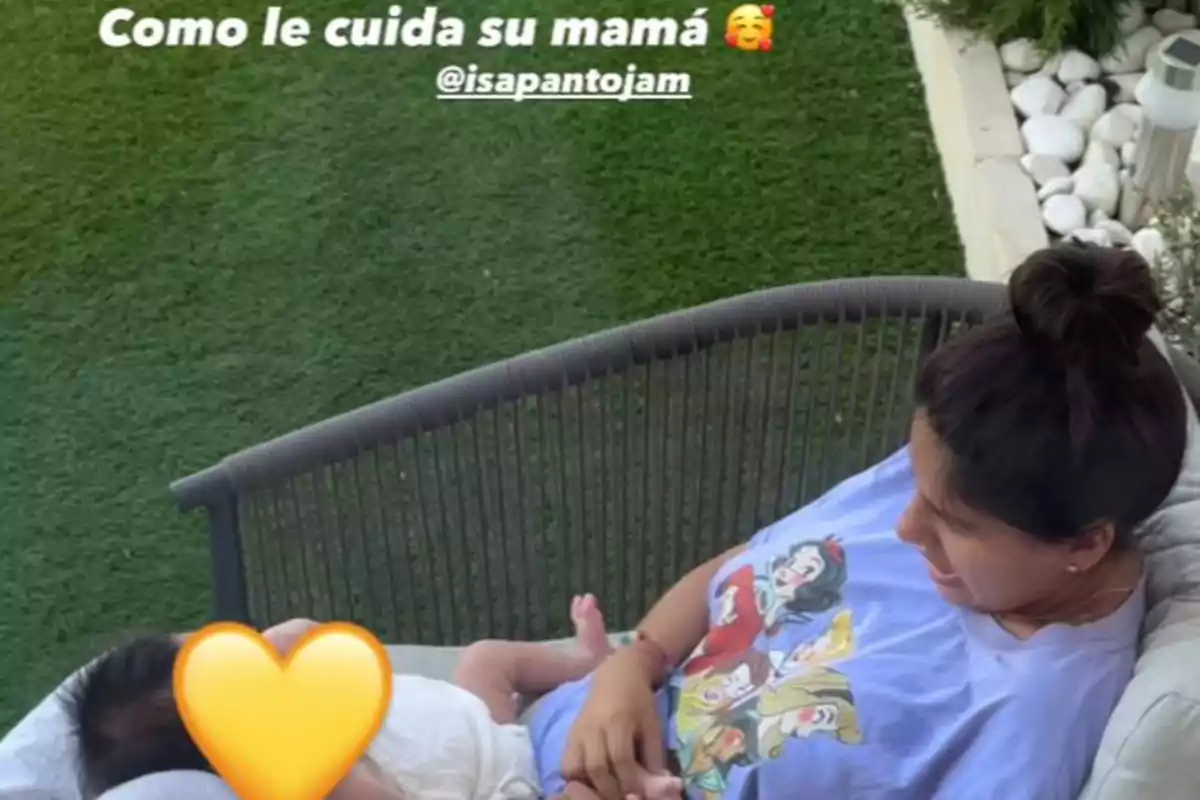 Isa Pantoja sentada en un sillón al aire libre sosteniendo a un bebé en brazos, con césped y piedras blancas de fondo, la mujer lleva una camiseta con princesas de Disney y el rostro del bebé está cubierto con un emoji de corazón amarillo Isa Pantoja sentada en un sillón al aire libre sosteniendo a un bebé en brazos, con césped y piedras blancas de fondo, la mujer lleva una camiseta con princesas de Disney y el rostro del bebé está cubierto con un emoji de corazón amarillo