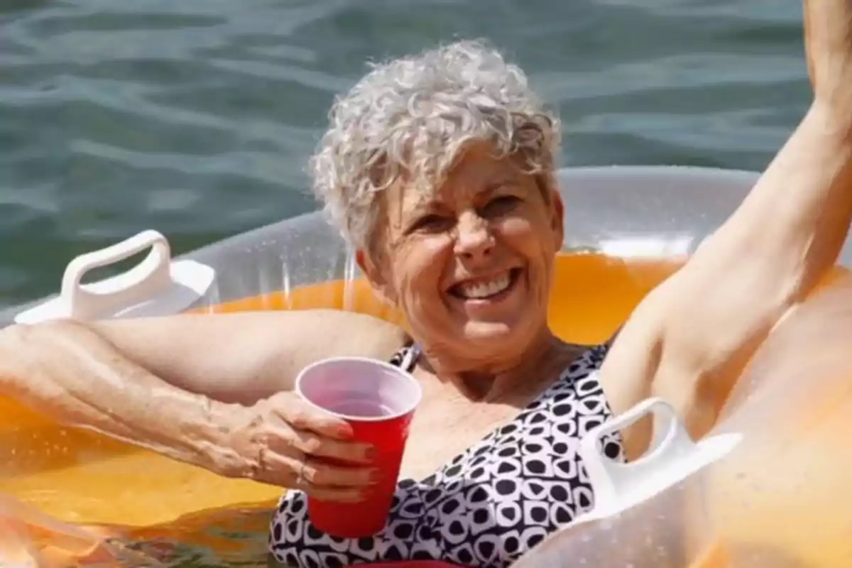 Dona gran somrient gaudint en una piscina sobre un inflable taronja mentre sosté un got vermell Dona gran somrient gaudint en una piscina sobre un inflable taronja mentre sosté un got vermell