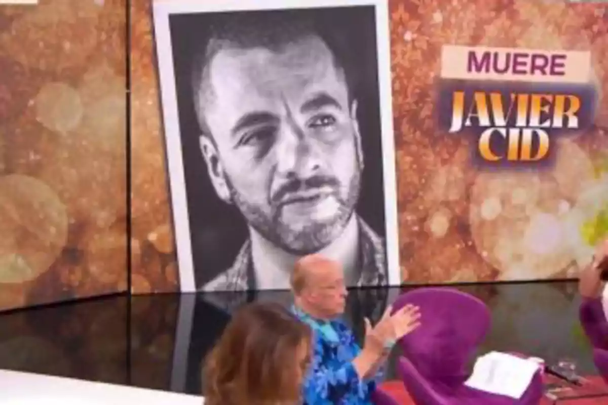 En un plató de televisió amb fons daurat es mostra una gran fotografia en blanc i negre d’un home i un rètol que diu mor Javier Cid mentre dues persones conversen en primer pla. En un plató de televisió amb fons daurat es mostra una gran fotografia en blanc i negre d’un home i un rètol que diu mor Javier Cid mentre dues persones conversen en primer pla.