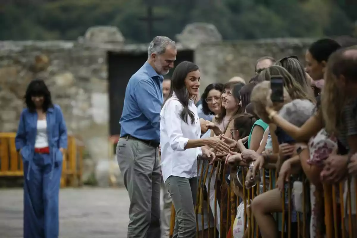 A couple greets and shakes hands with several people who are behind a fence in an outdoor setting with a stone wall in the background. A couple greets and shakes hands with several people who are behind a fence in an outdoor setting with a stone wall in the background.