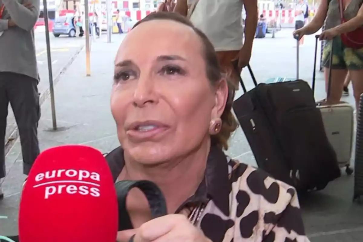Benita siendo entrevistada en la calle con un micrófono rojo de europa press y maletas de viaje al fondo