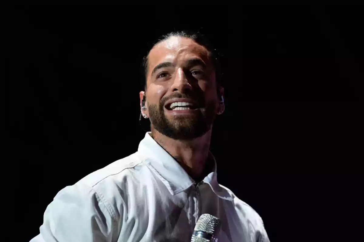 Bearded man with tied-back hair smiles while holding a microphone on stage with a dark background Bearded man with tied-back hair smiles while holding a microphone on stage with a dark background