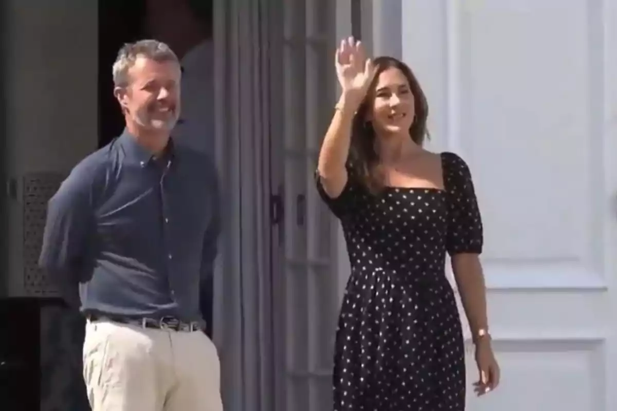 A woman in a black polka-dot dress waves while a man in a blue jersey and light-colored pants smiles next to her in front of a white door. A woman in a black polka-dot dress waves while a man in a blue jersey and light-colored pants smiles next to her in front of a white door.