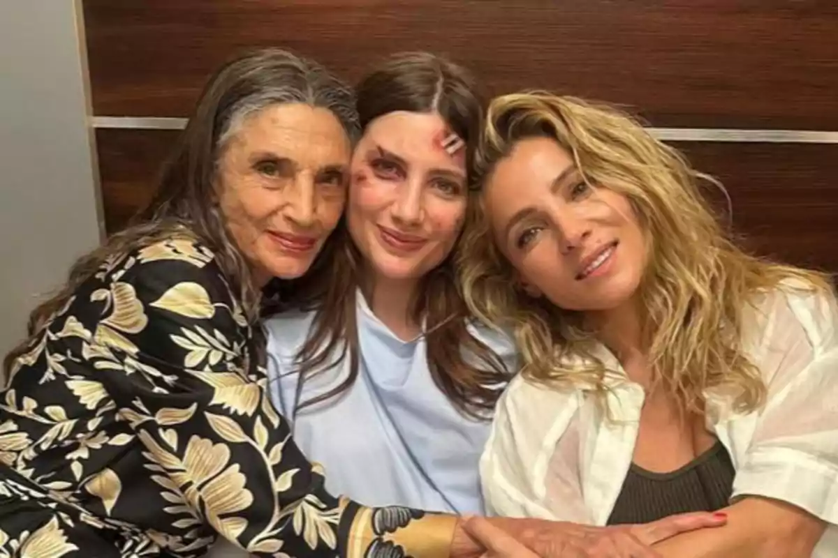 Tres mujeres sentadas abrazándose y sonriendo, la del centro tiene heridas simuladas en la cara y lleva una bata azul clara Tres mujeres sentadas abrazándose y sonriendo, la del centro tiene heridas simuladas en la cara y lleva una bata azul clara
