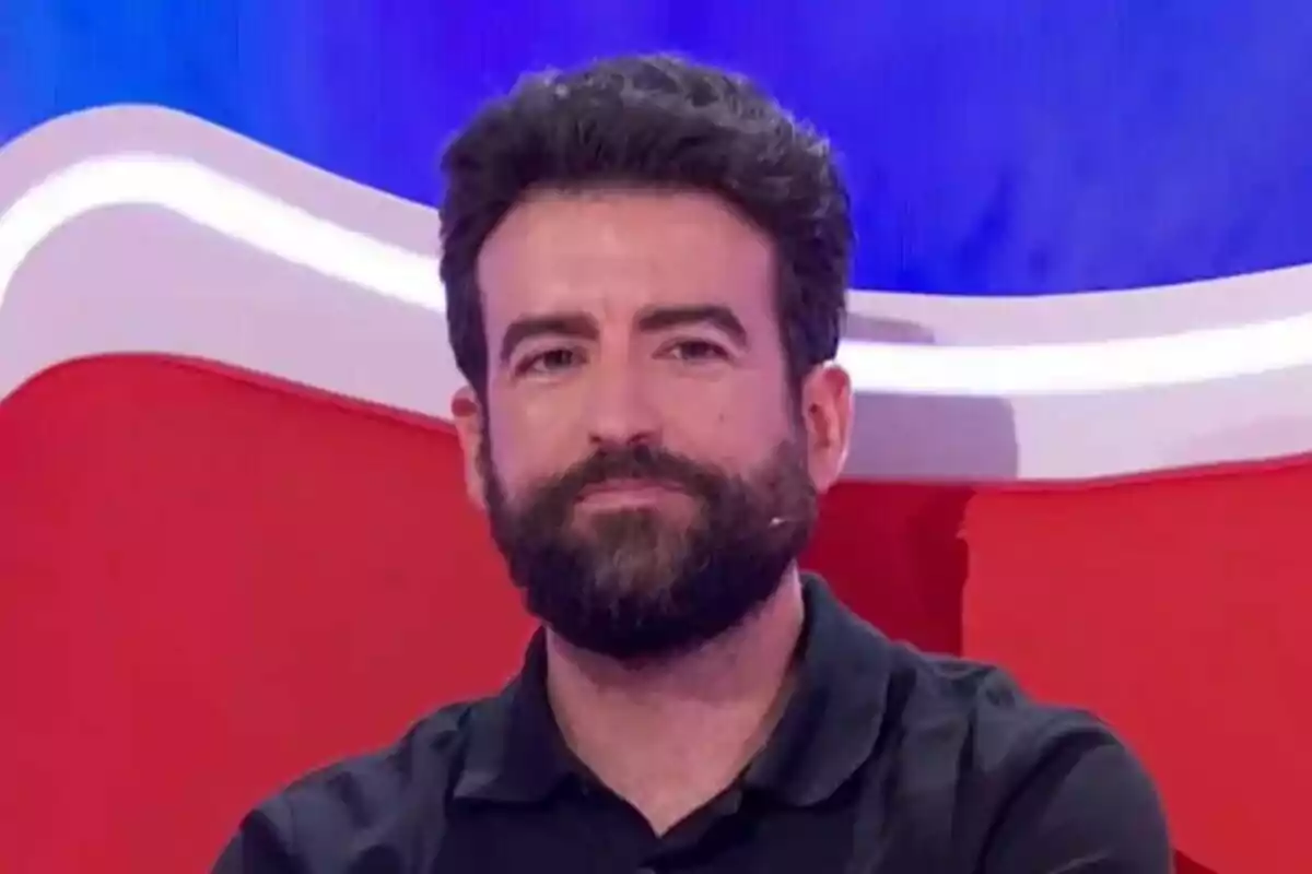 Plano corto de Rafa Castaño con barba y cabello oscuro sentado en un sillón rojo con fondo azul, que es el plató de 'Agárrate al Sillón'. Plano corto de Rafa Castaño con barba y cabello oscuro sentado en un sillón rojo con fondo azul, que es el plató de 'Agárrate al Sillón'.