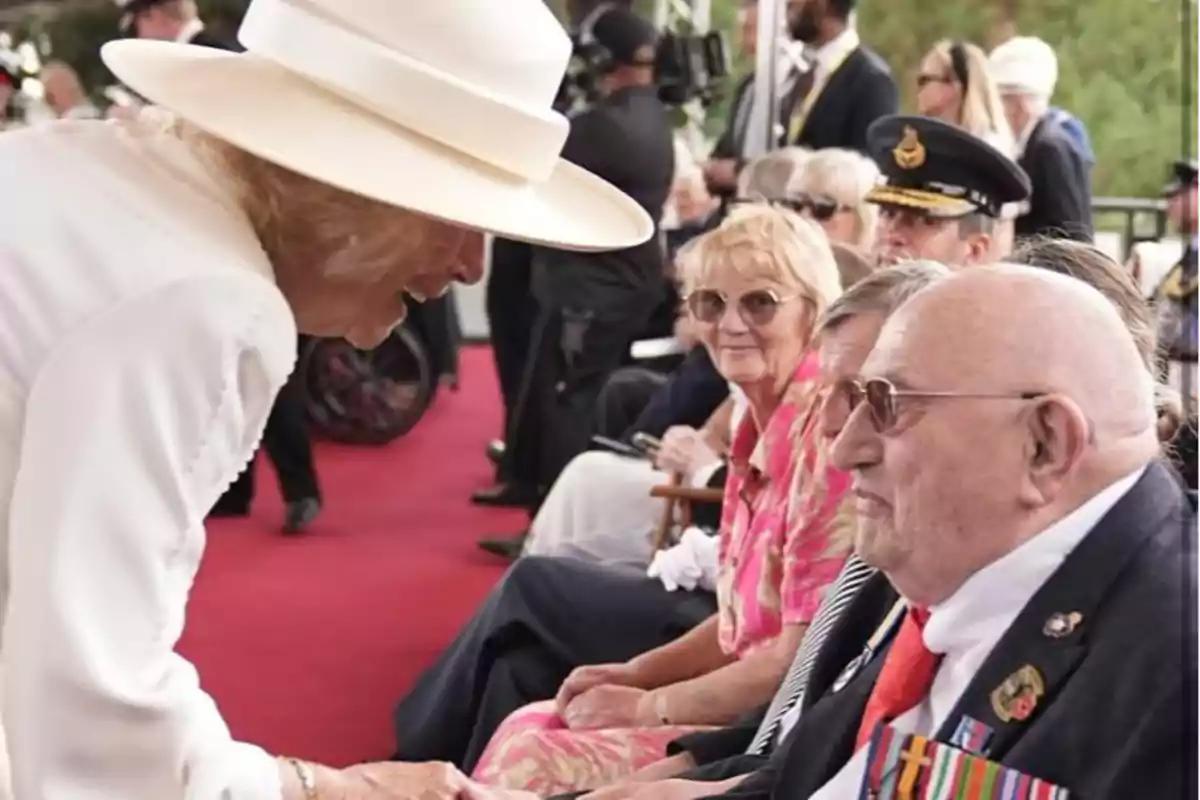 La reina Camila vestida de blanco y con sombrero saluda a un hombre mayor con medallas en el pecho que está sentado junto a otras personas en un evento formal