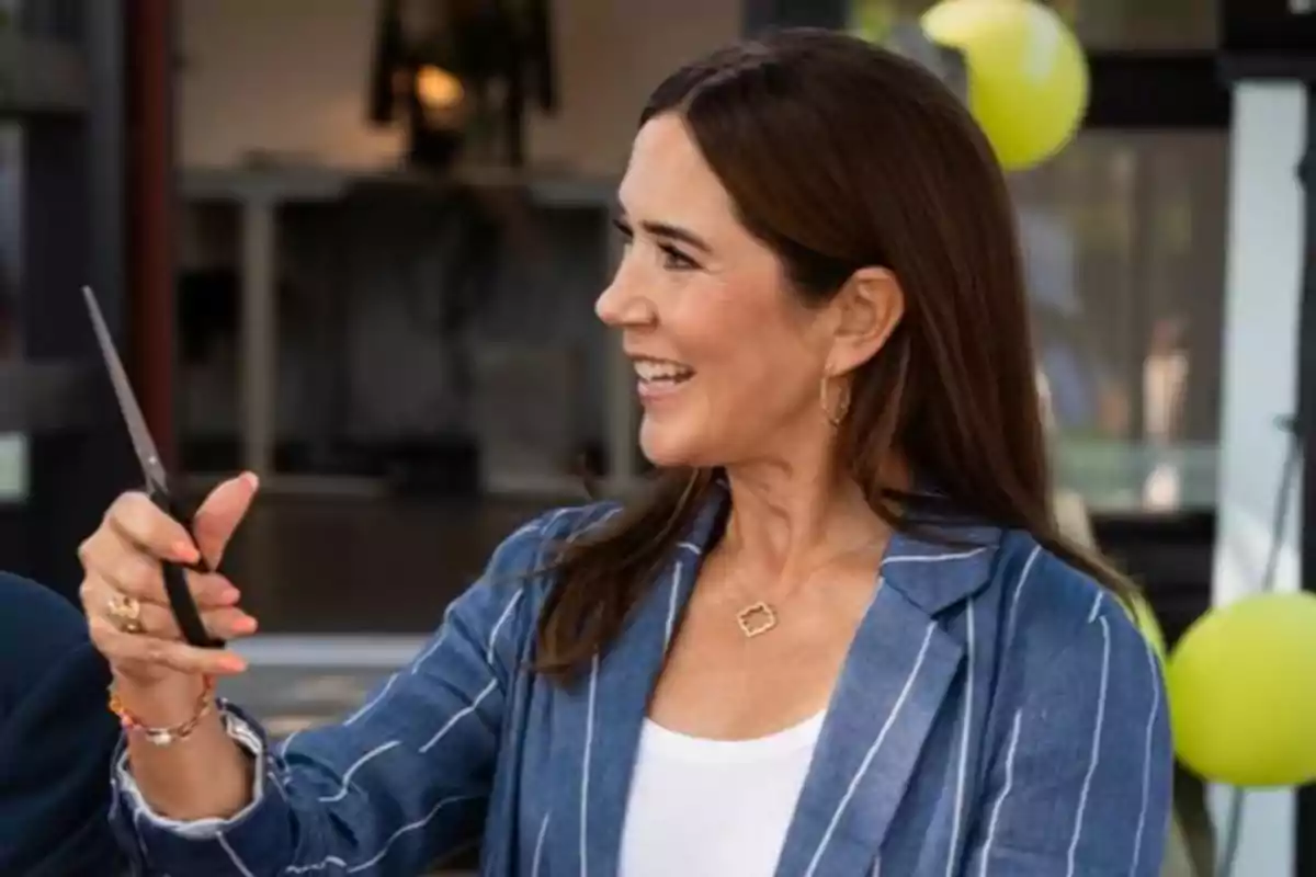 La reina Mary sonriente con tijeras en la mano vestida con saco azul y camiseta blanca en un evento con globos amarillos al fondo La reina Mary sonriente con tijeras en la mano vestida con saco azul y camiseta blanca en un evento con globos amarillos al fondo