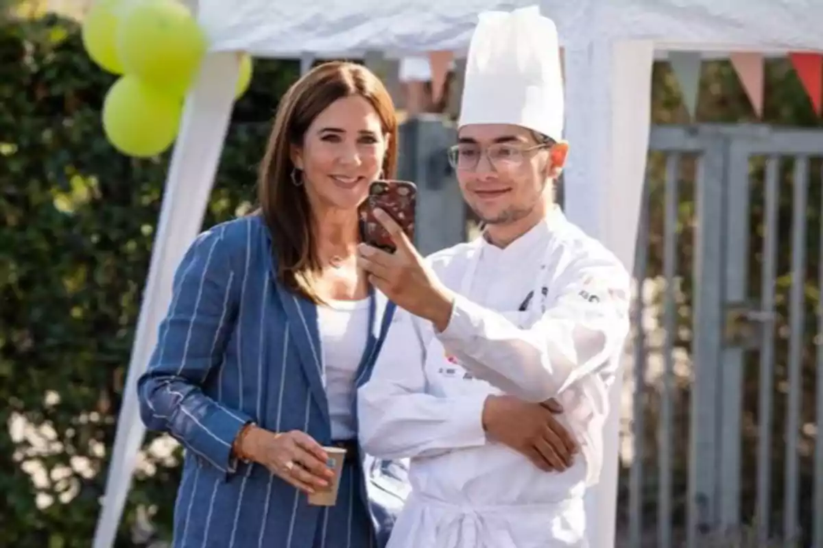 La reina Mary y un chef sonríen mientras se toman una selfie al aire libre en un evento con globos y decoración festiva La reina Mary y un chef sonríen mientras se toman una selfie al aire libre en un evento con globos y decoración festiva