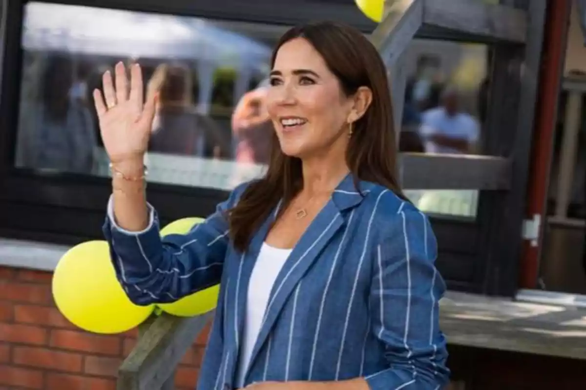 La reina Mary sonriente con chaqueta azul de rayas saludando con la mano frente a globos amarillos La reina Mary sonriente con chaqueta azul de rayas saludando con la mano frente a globos amarillos